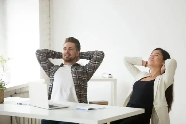 A man and woman sitting at a table with their hands behind their head.