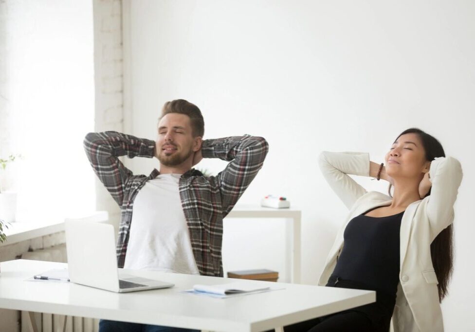 A man and woman sitting at a table with their hands behind their head.